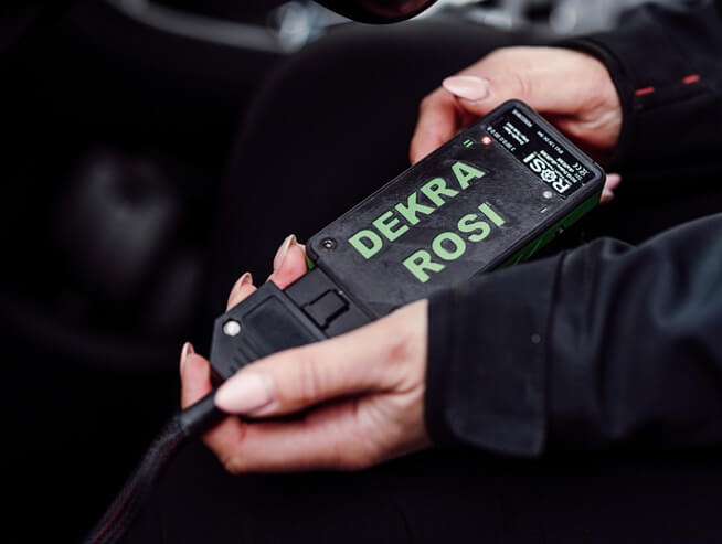 Närbild på två händer som håller i ett svart diagnostikverktyg med den gröna texten ”DEKRA ROSI”. Verktyget är kopplat till en kabel.