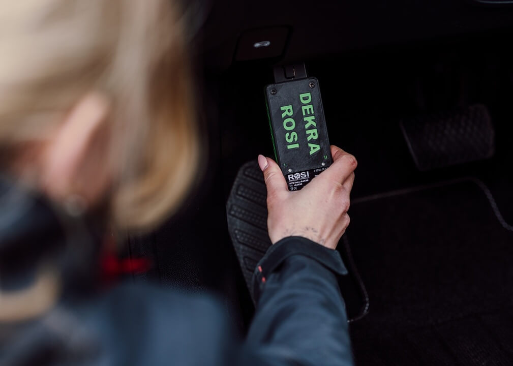Person håller en diagnostikenhet märkt ‘DEKRA ROSI’ ansluten till ett fordons uttag för analys och kontroll av bilens system.